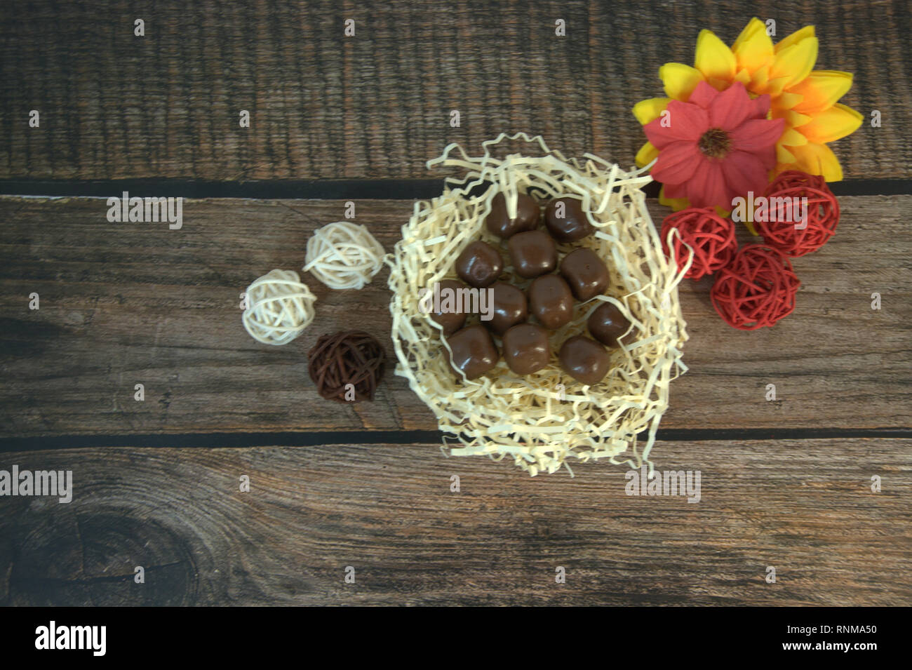 Round chocolates in a nest of straw, multicolored decorative balls and flower buds on a wooden table. Close-up. Stock Photo