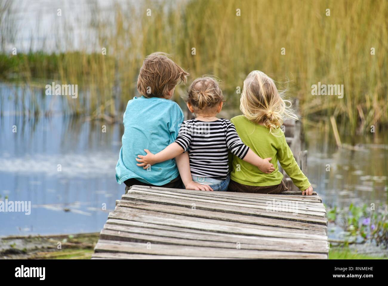 Three toddlers sitting together on an old jetty, back view, Patagonia, Argentina Stock Photo