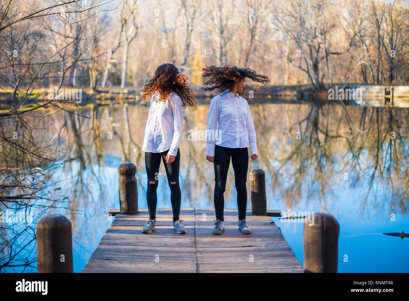 Young twin sisters in same clothes shaking hair at lake on autumn day ...