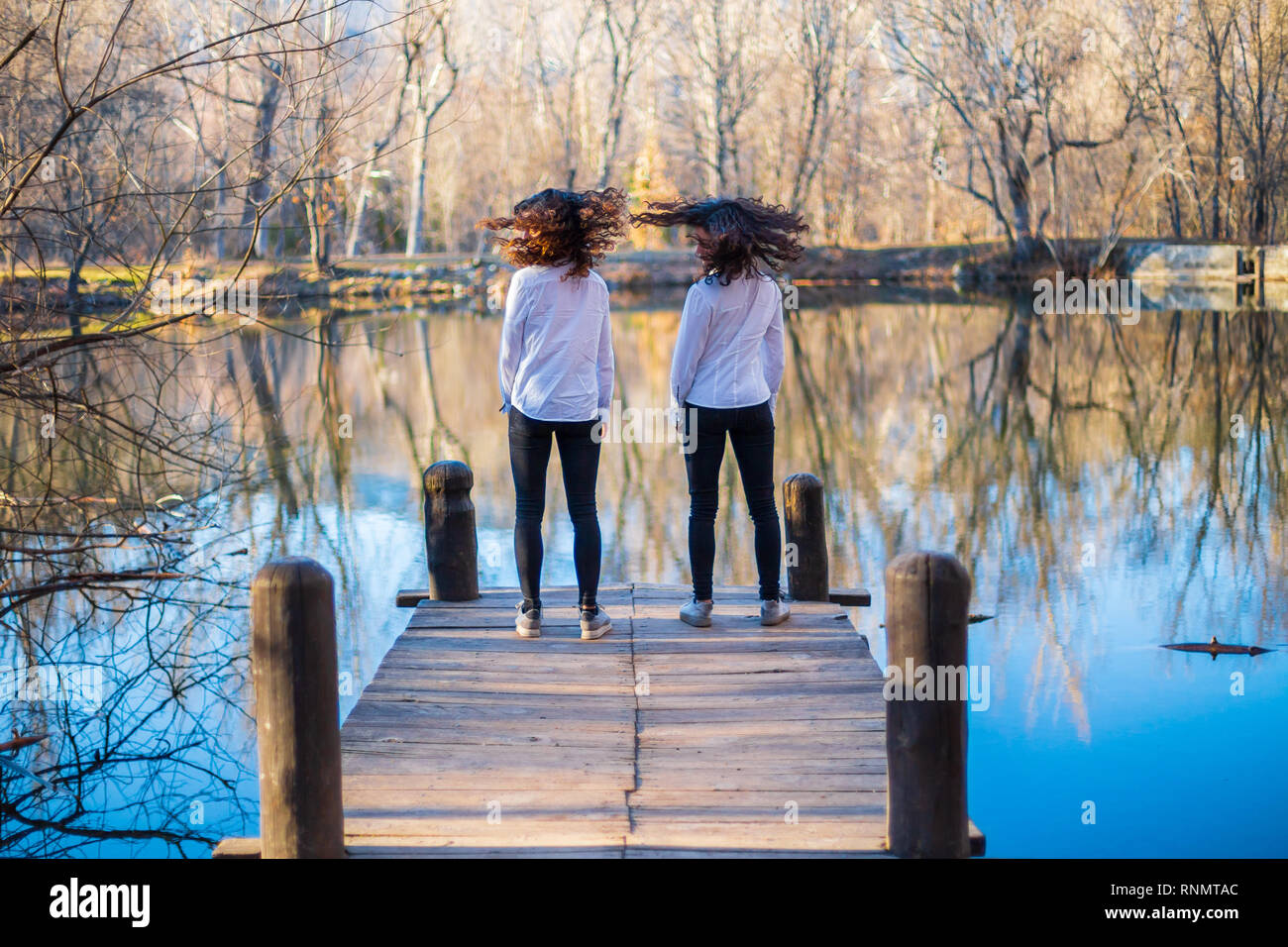 Back view of young twin sisters in same clothes shaking hair at lake on autumn day in the forest Stock Photo