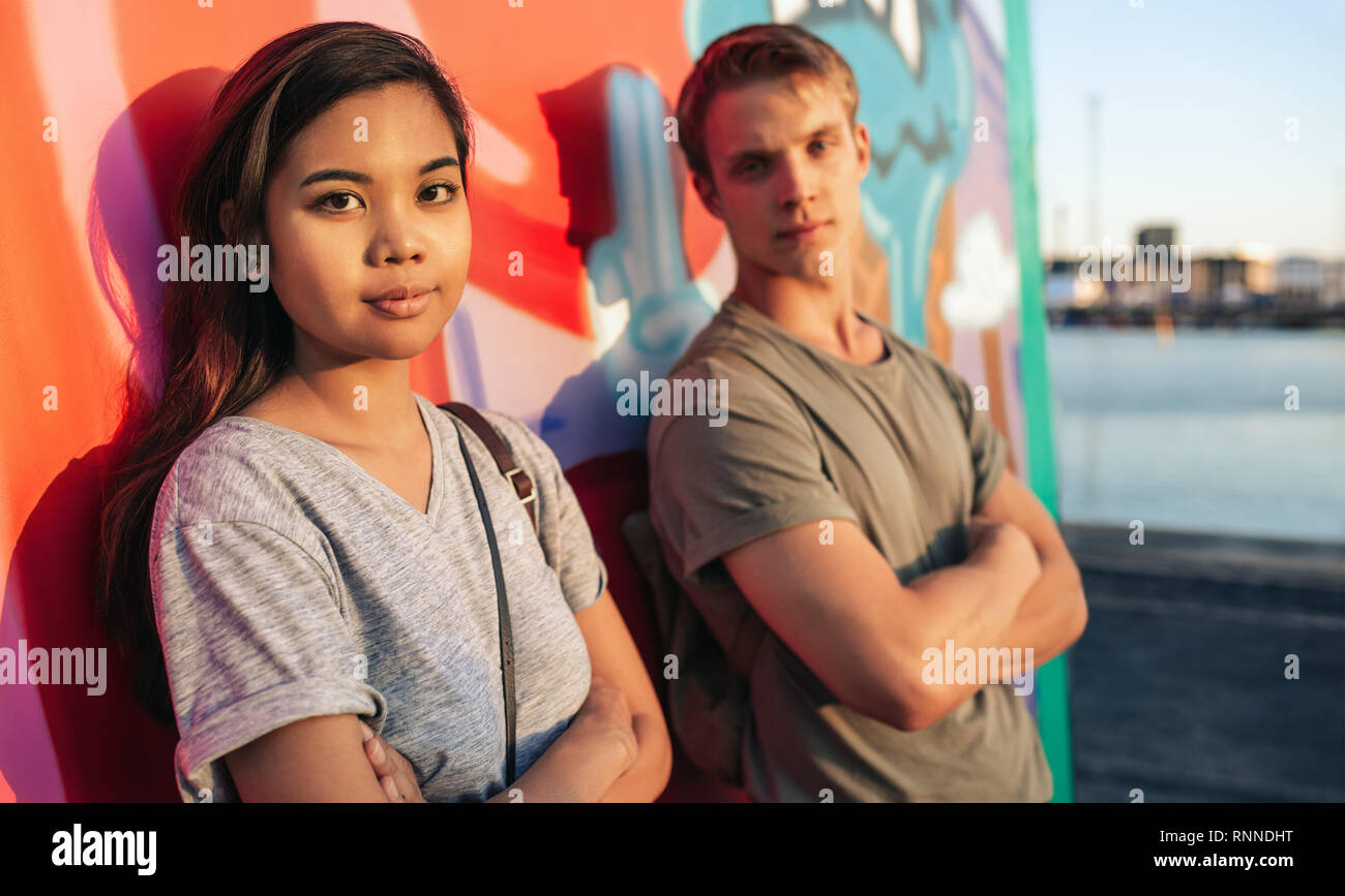 Young woman with attitude leaning with her arms crossed on a wall of graffiti in the city with her boyfriend in the background Stock Photo