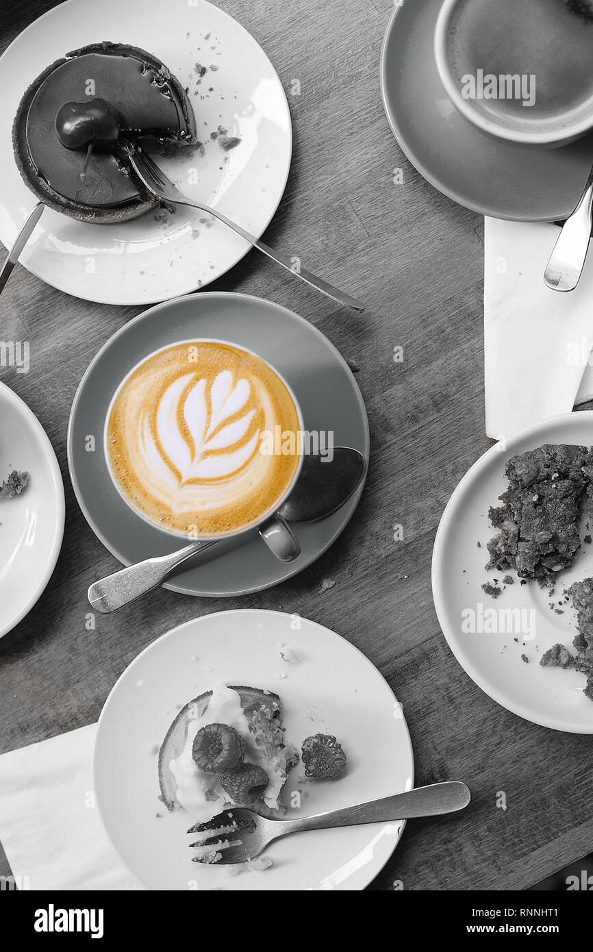 Dining table filled with half eaten food with a full cup of coffee untouched and decorated with foam latte art. Photo in monochrome and color. Stock Photo