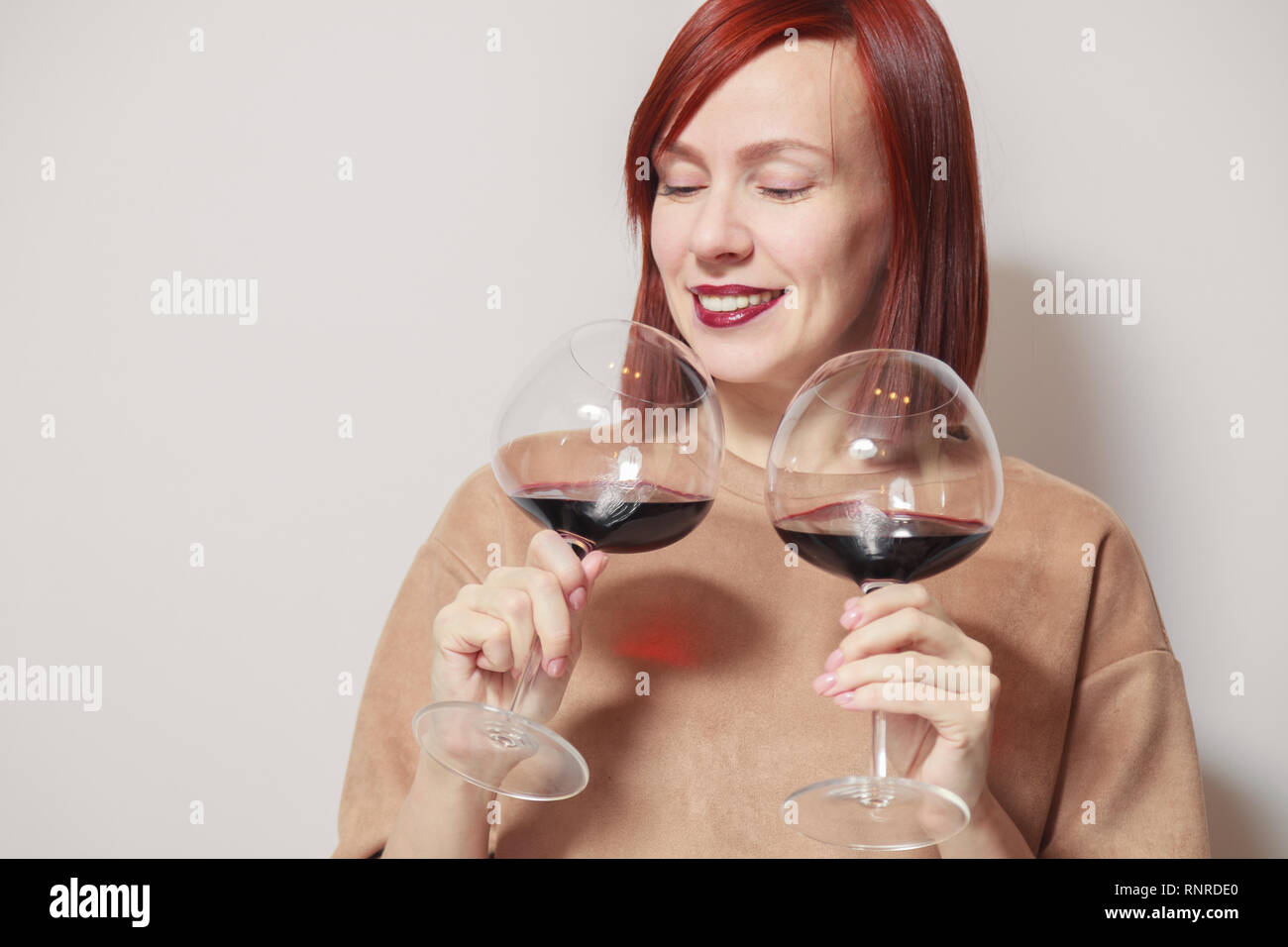 Young cute beautiful redhaired girl with bright lipstick on a white background holds and sniffs two glasses of red wine and assesses the aroma and not Stock Photo