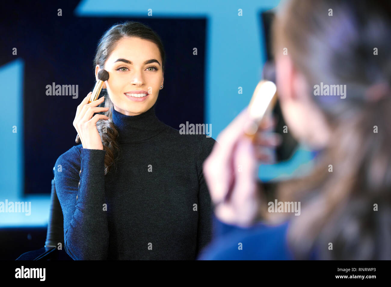 Portrait of gorgeous young woman applying her makeup while sitting at mirror in the photostudio. Stock Photo