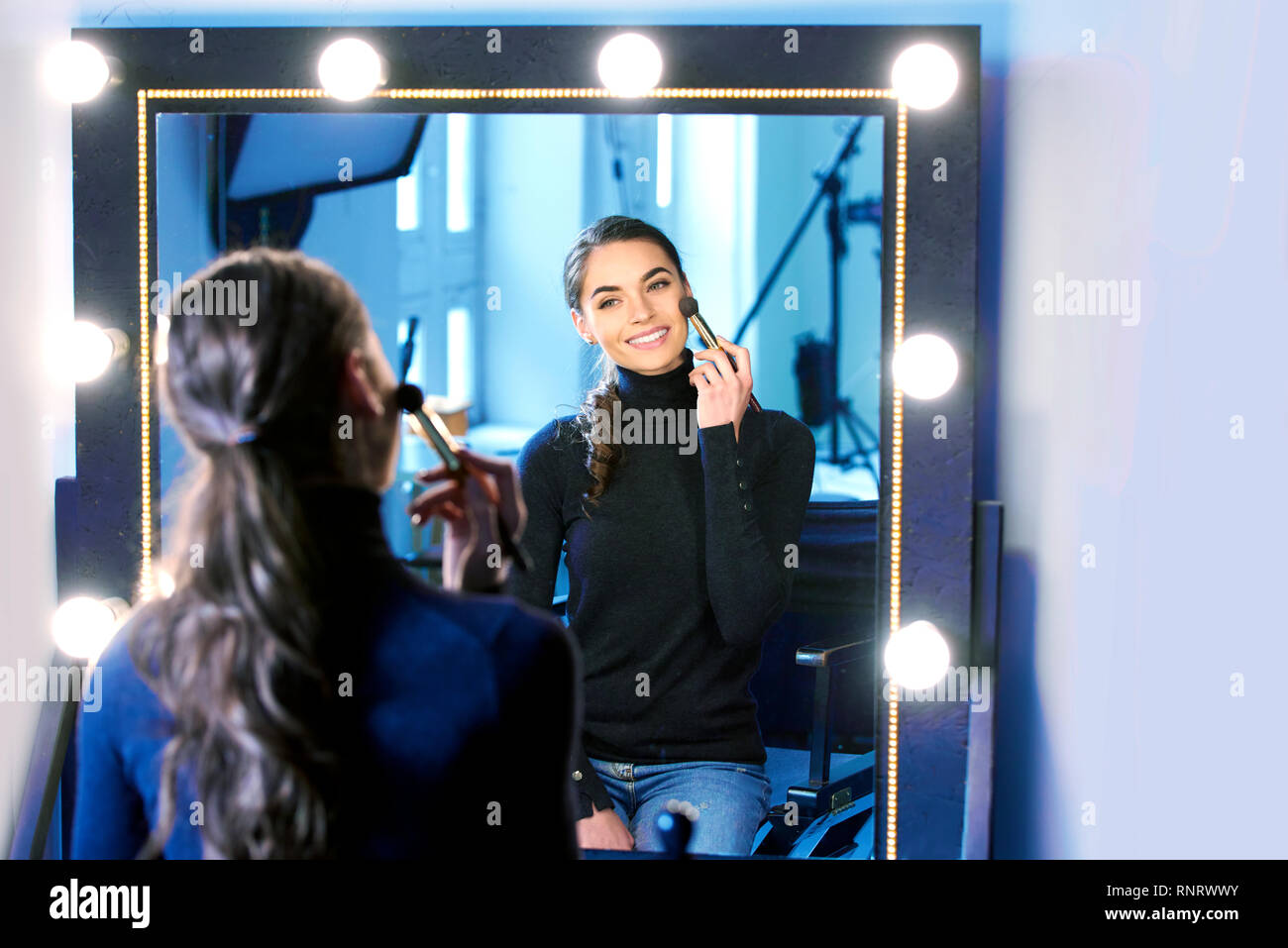 Portrait of gorgeous young woman applying her makeup while sitting at mirror in the photostudio. Stock Photo