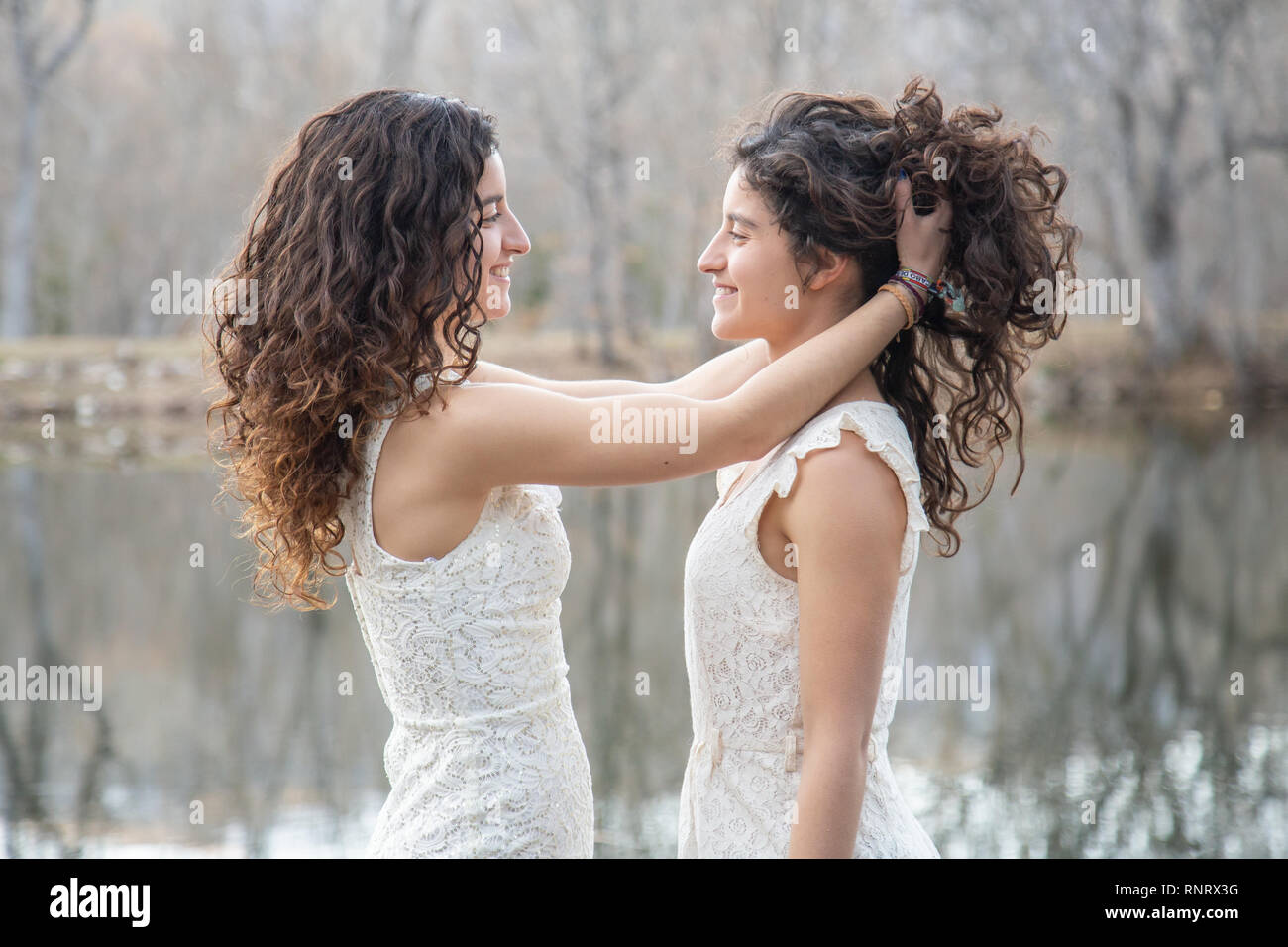 Side view of cheerful twin sisters standing at calm lake smiling and combing hair to each other on autumn day Stock Photo