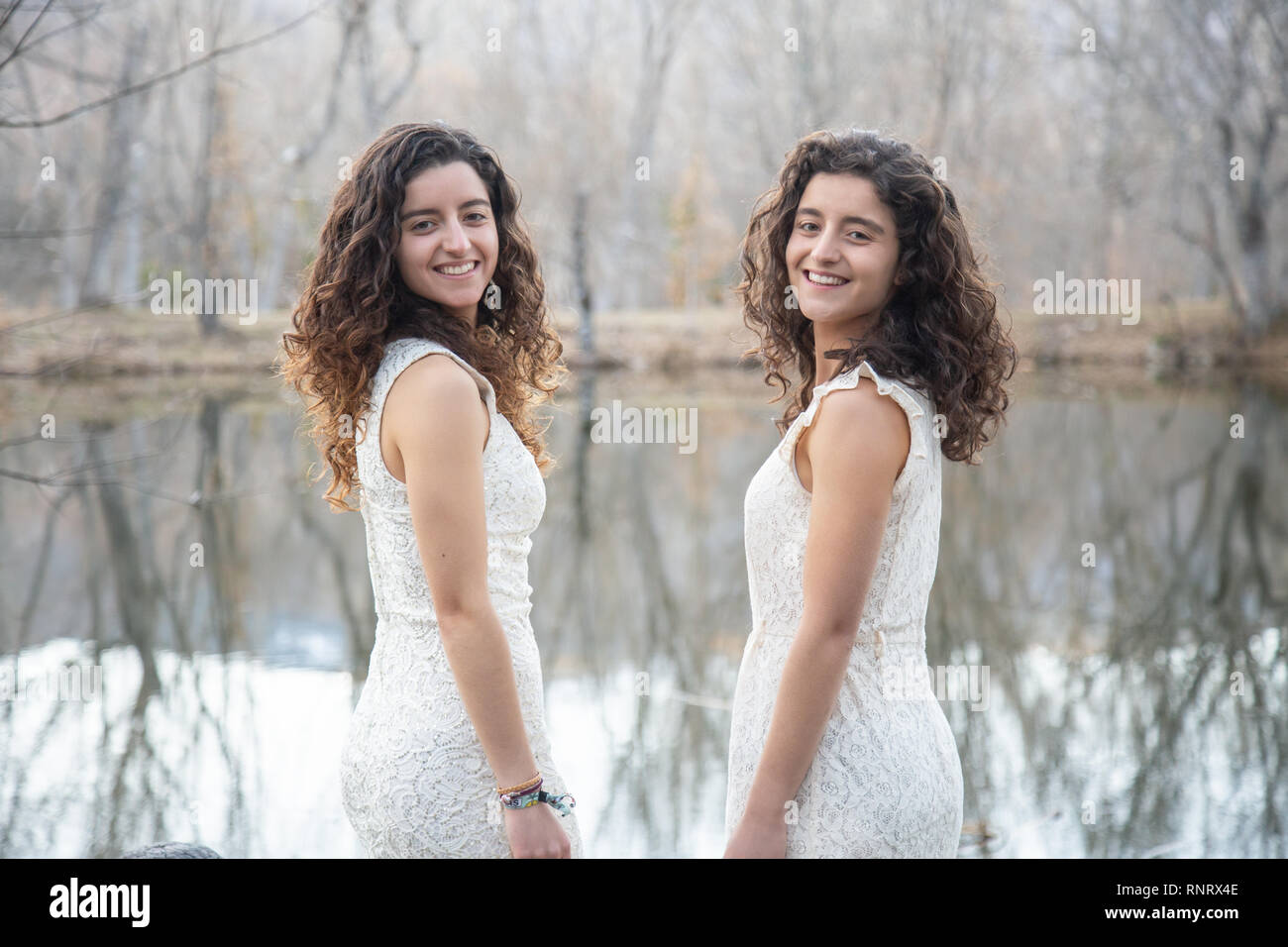 Side view of cheerful twin sisters standing at calm lake smiling and ...