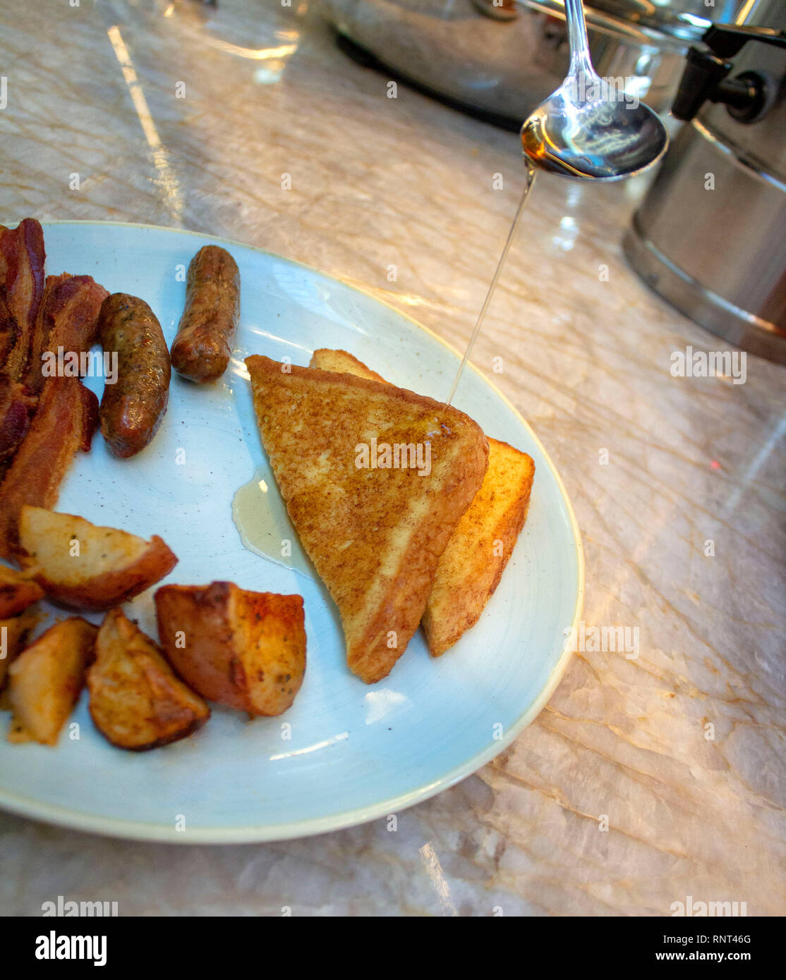 Syrup being poured on french toast on a plate with bacon, sausage links, and potatoes. Stock Photo