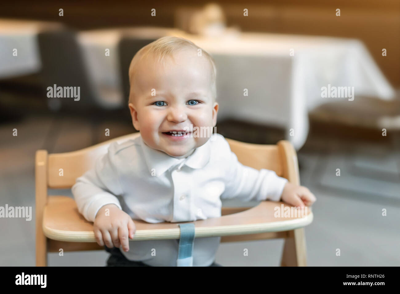 Cute little baby boy in white polo tshirt sitting in wooden baby chair and laughing at cafe