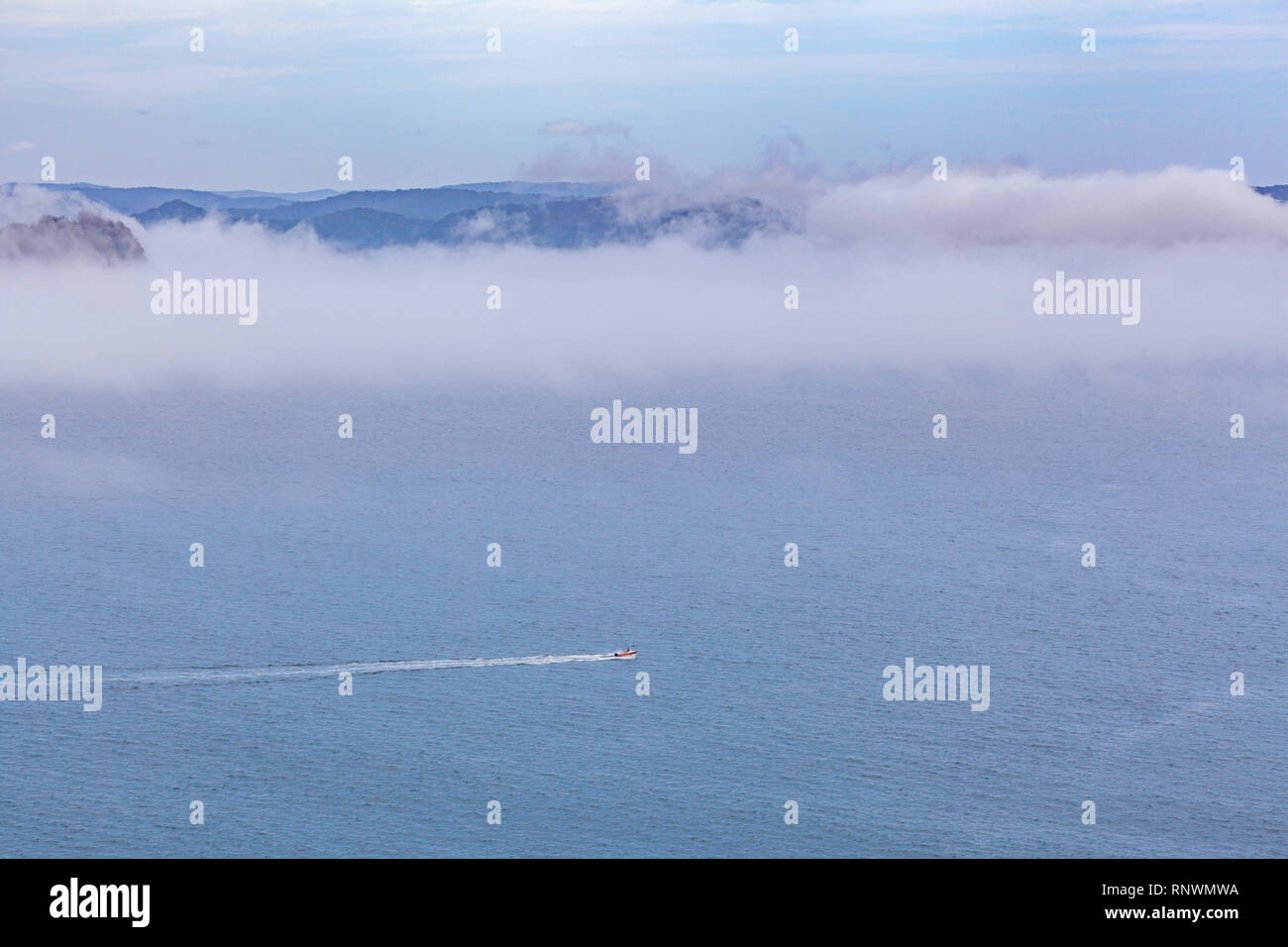 Small boat sailing across the ocean leaving water trail under low clouds Stock Photo