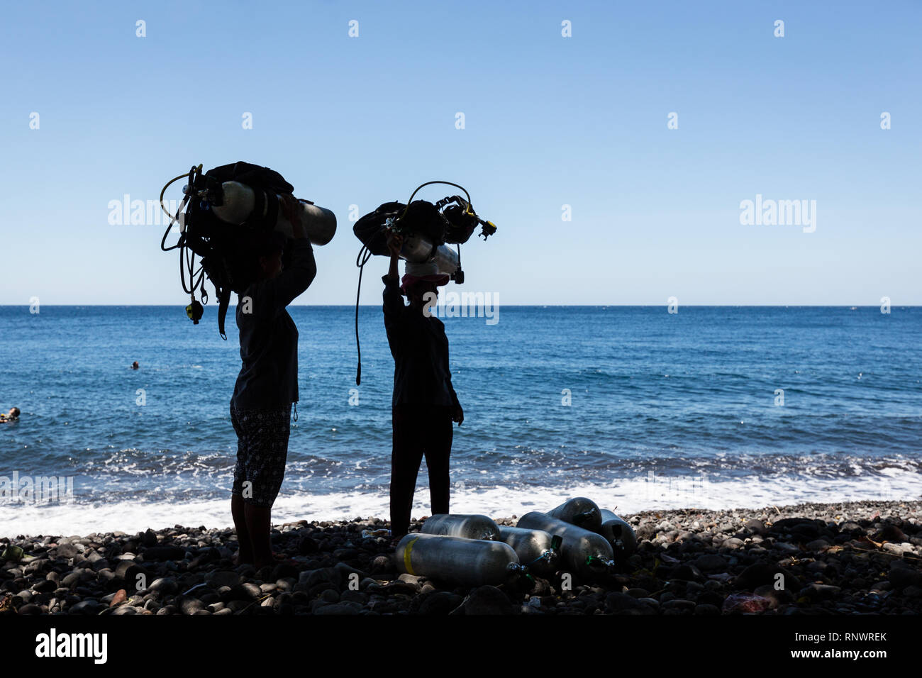 Two women carry heavy scuba diving gear on their heads, a common site in Tulemban, Bali, Indonesia. Stock Photo