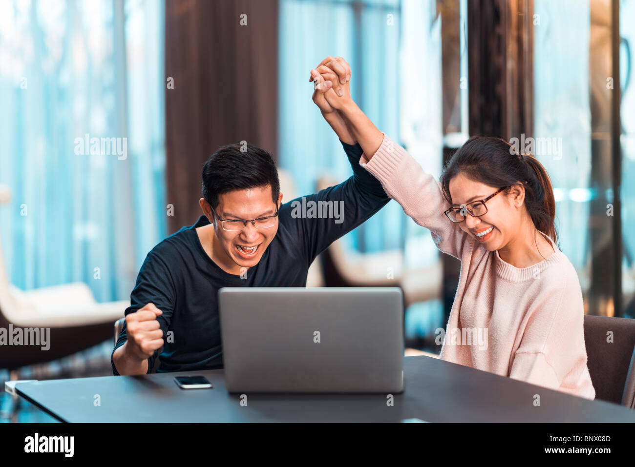 Young Asian married couple hold hands celebrating together, watching laptop computer in modern home office. Success job, love relationship concept Stock Photo