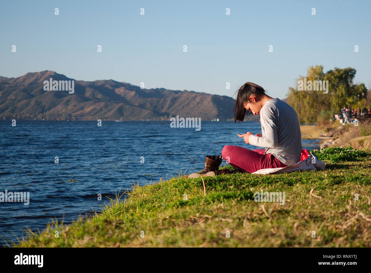Villa Carlos Paz, Cordoba, Argentina - 2019: A young woman rests with her shoes off  by the San Roque lake on a Sunday evening. Stock Photo