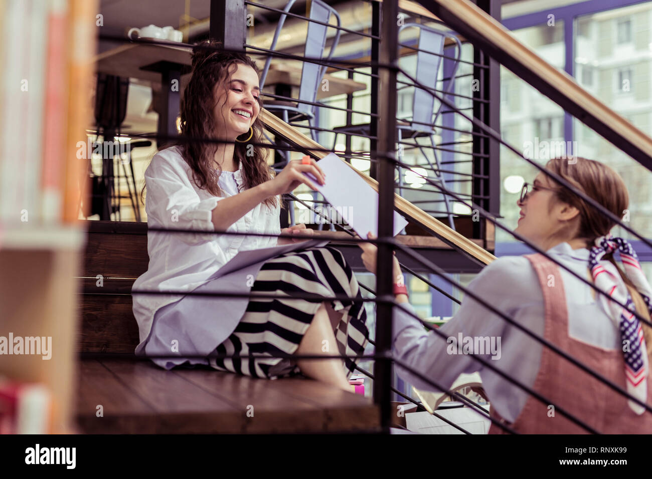 Positive young girls exchanging papers while sitting on metal stairs Stock Photo