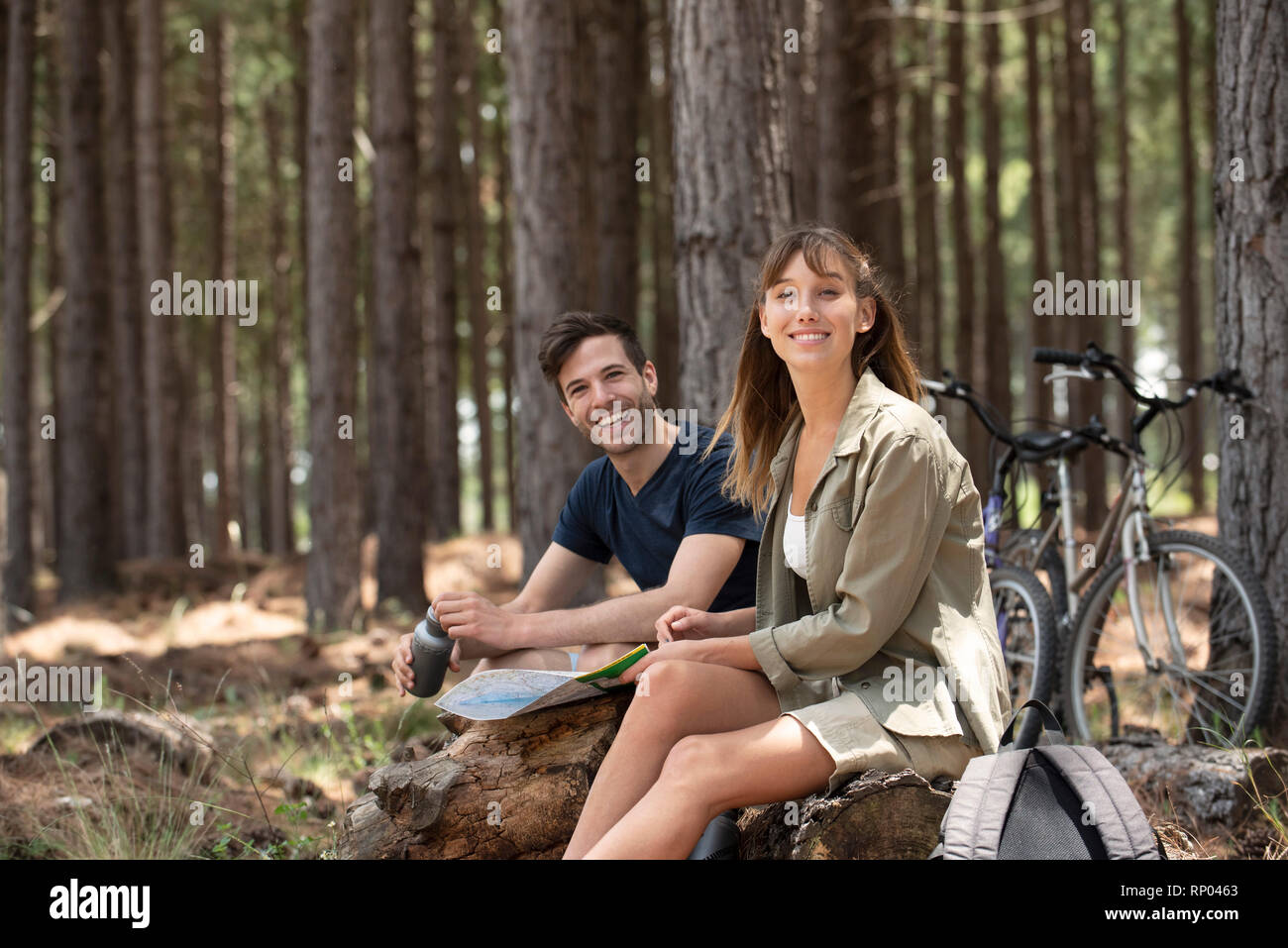 Smiling young couple sitting on log in pine forest Stock Photo