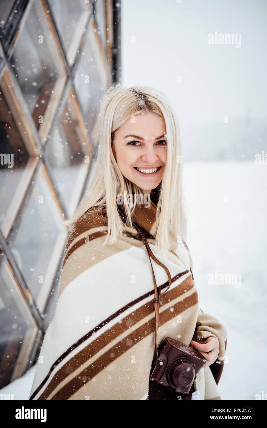 Beautiful blonde girl with a vintage camera smiling against the backdrop of a winter landscape. Nikola-Lenivets, Russia Stock Photo