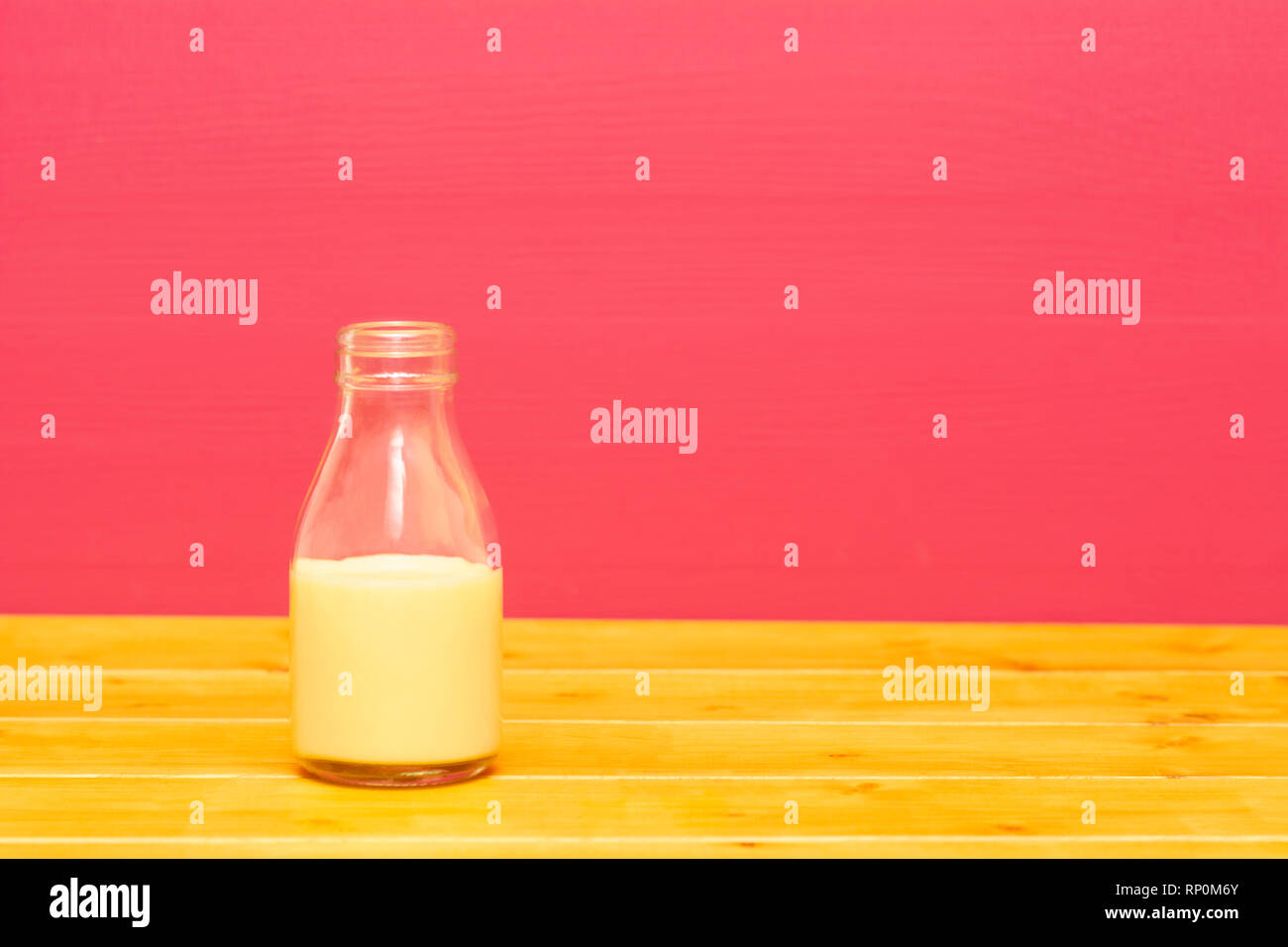 One-third pint glass milk bottle half full with banana milkshake, on a wooden table against a pink painted background Stock Photo