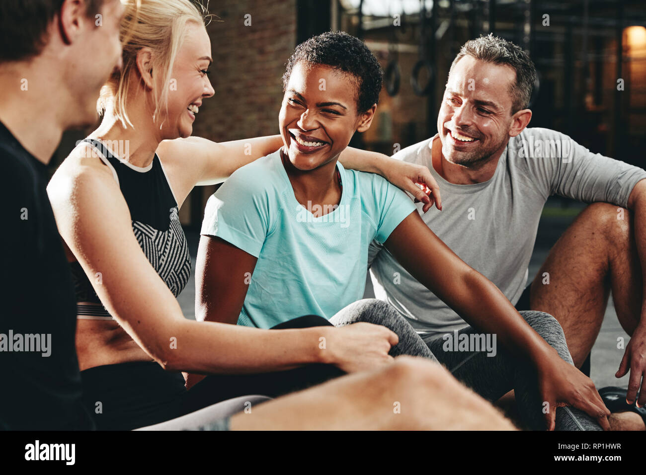 Smiling group of diverse people in sportswear sitting on a gym floor talking and laughing together after a workout class Stock Photo