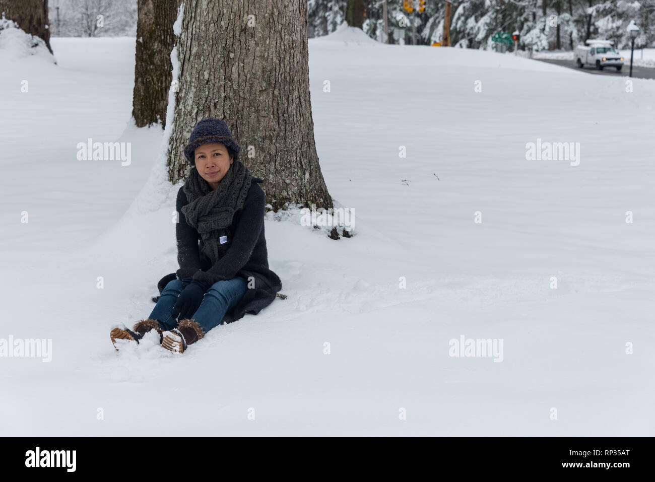 Asian woman in hat sitting in snow in front of tree Stock Photo