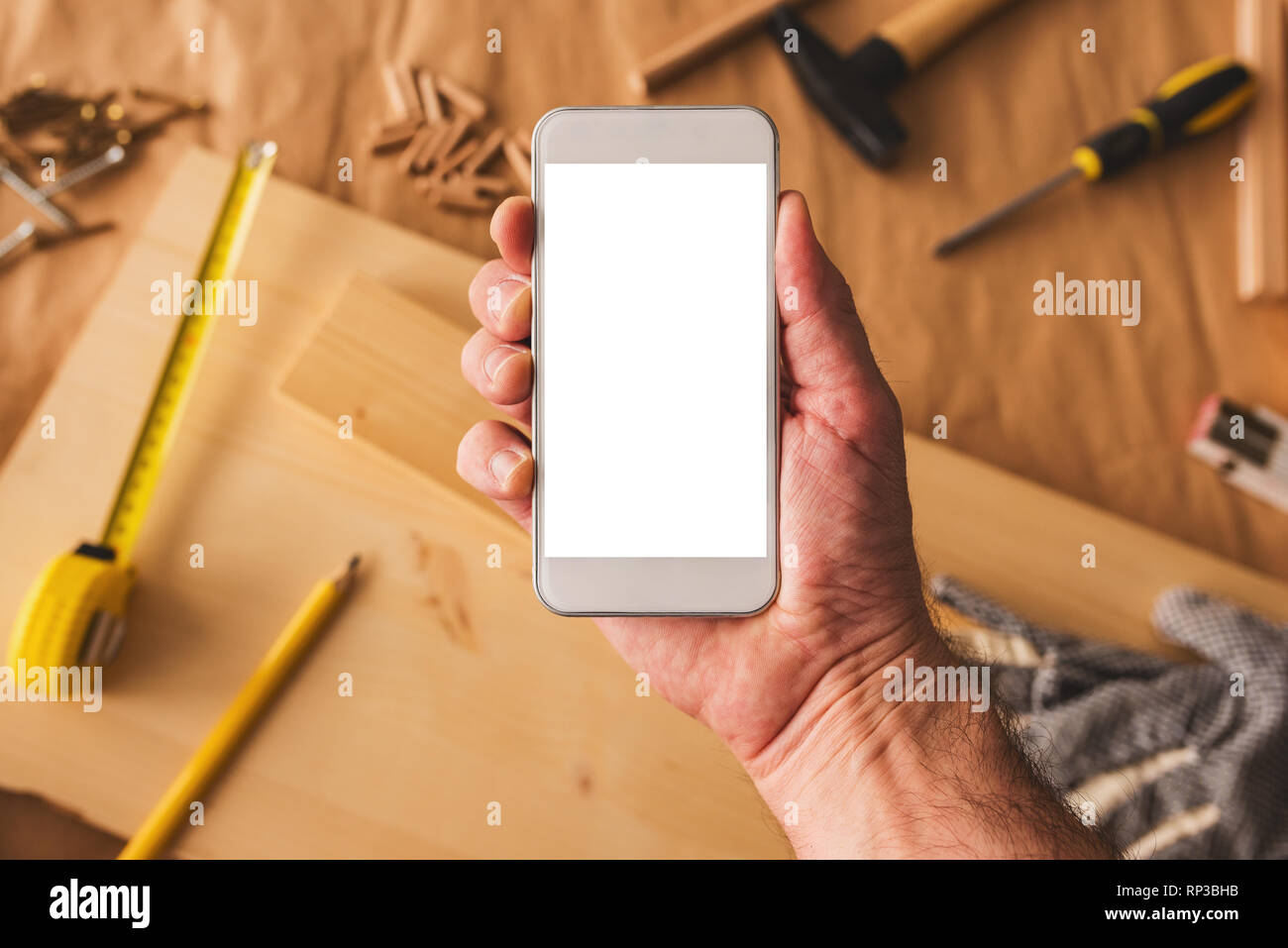 Small business woodwork entrepreneur holding modern smartphone device with blank mock up screen over workshop desk with tools Stock Photo