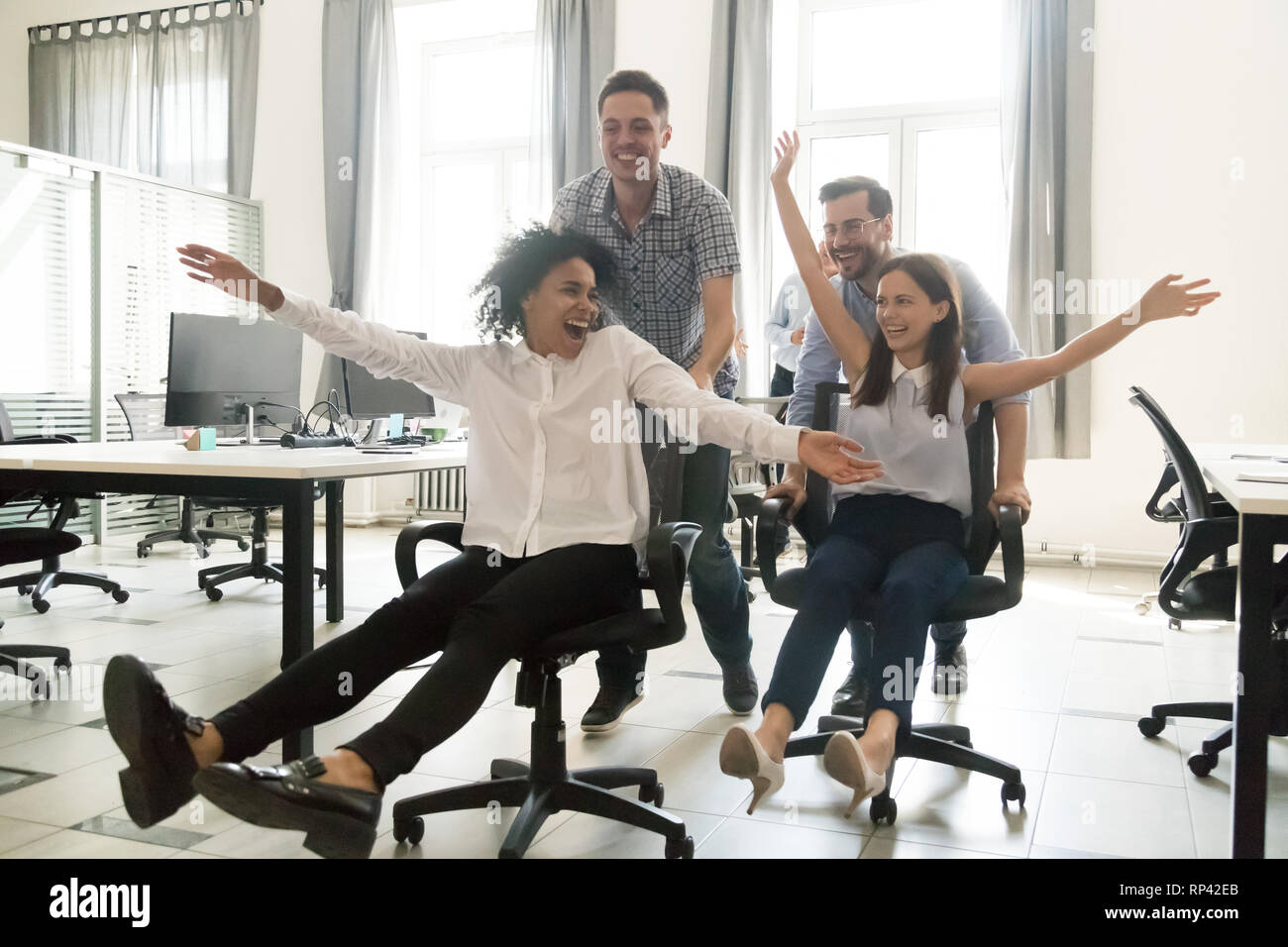 Excited young cheerful multiracial diverse colleagues having fun riding on office chairs in coworking space. Active happy millennial workers take a br Stock Photo