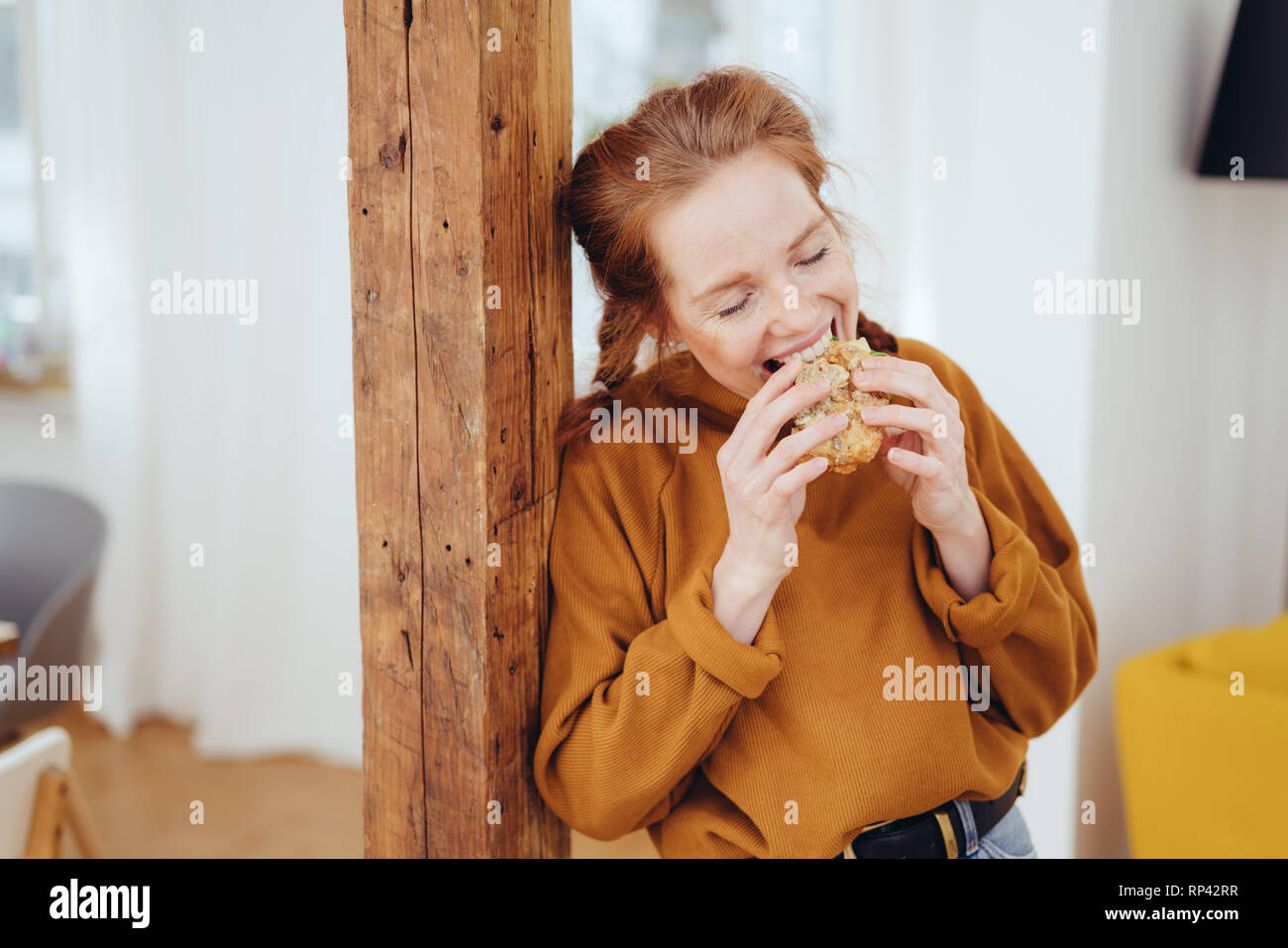 Hungry young woman taking a bite of a healthy wholewheat sandwich with a happy smile leaning on a wooden pillar indoors at home Stock Photo
