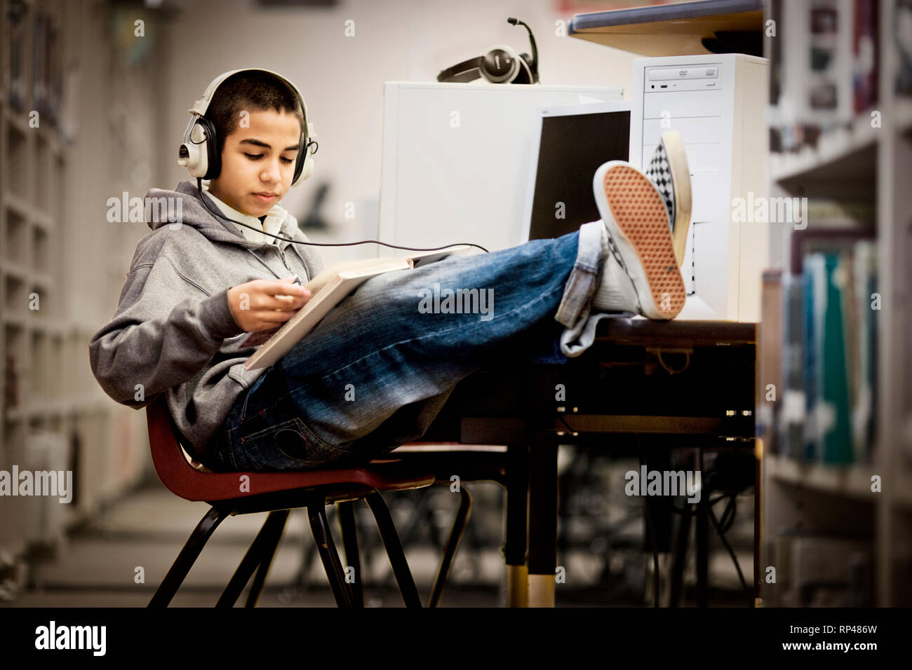 Teenage boy sitting with his feet up and reading a book with headphones on in a library. Stock Photo