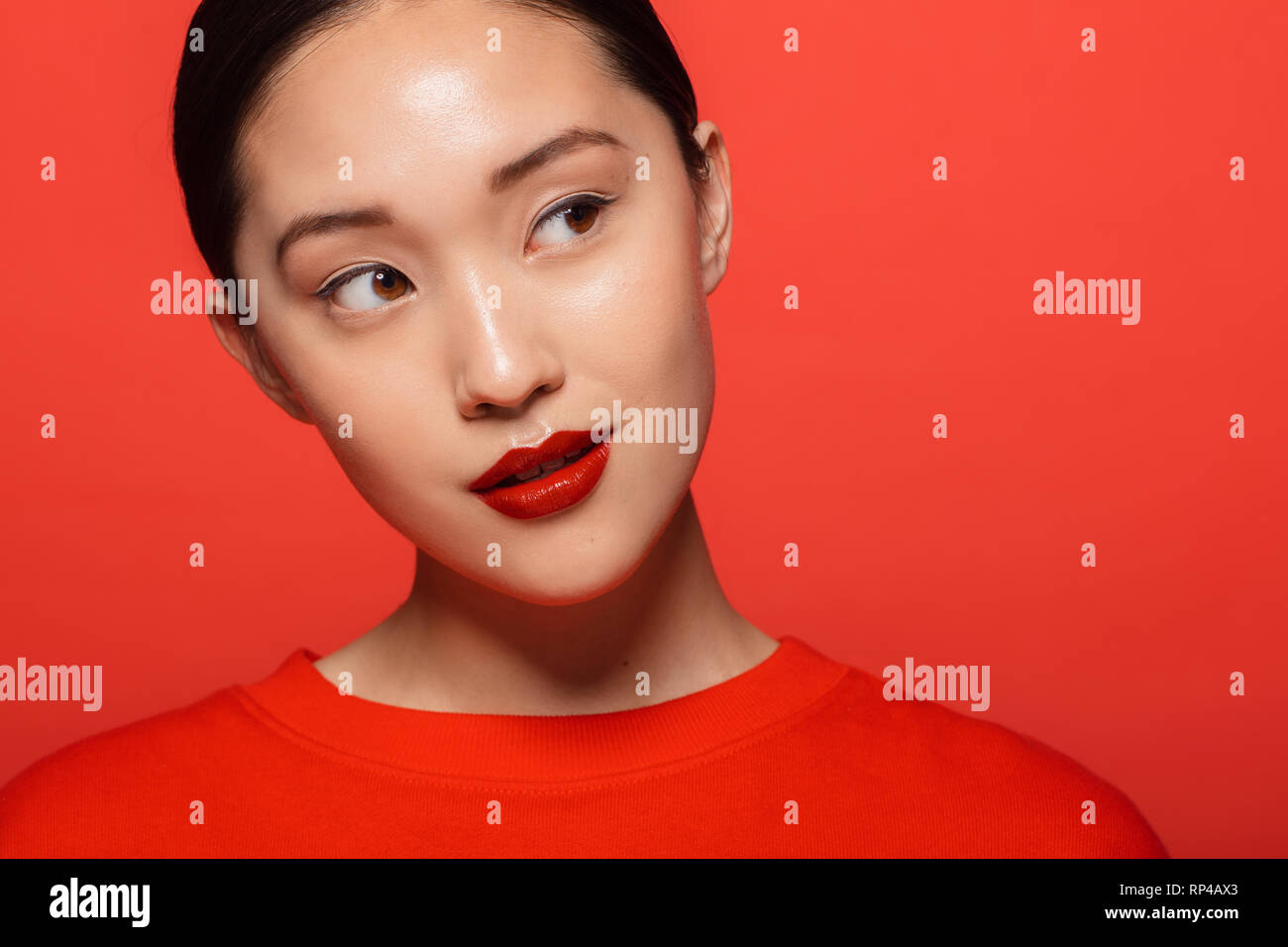 Close up of young asian woman with beautiful make up looking away and thinking. Korean female model with red make up against red background. Stock Photo