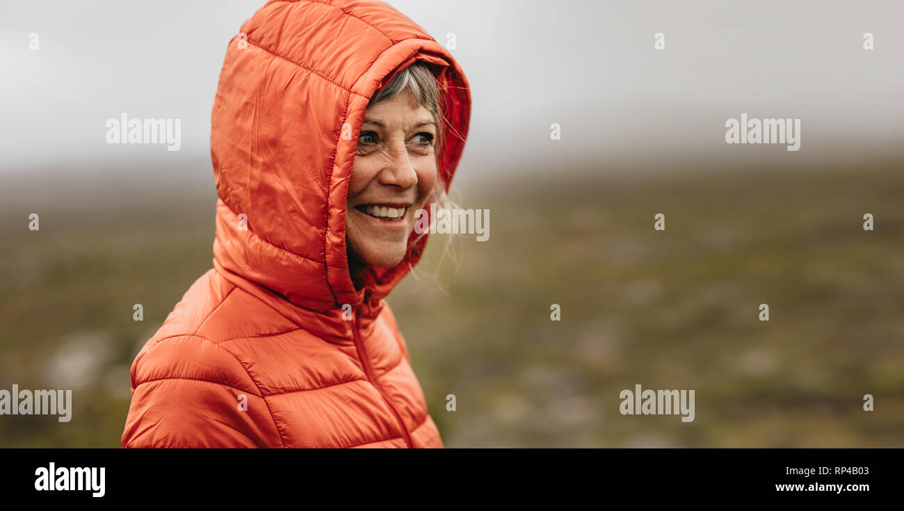 Smiling woman in a warm hooded jacket standing outdoors. Cheerful senior woman standing on a hill looking away. Stock Photo