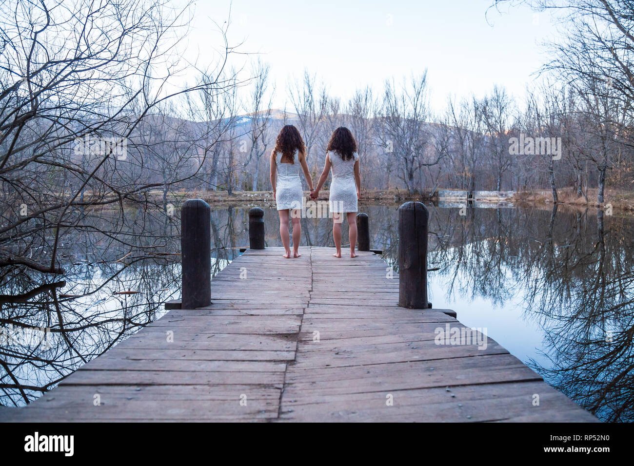 Back view of twin sisters in same white dresses holding hands while standing on wooden pier at calm lake on autumn day Stock Photo