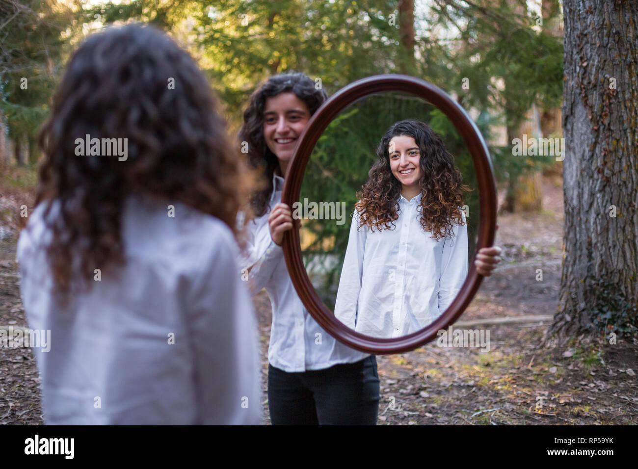 Cheerful young woman showing reflection of twin sister in autumn day in the forest Stock Photo