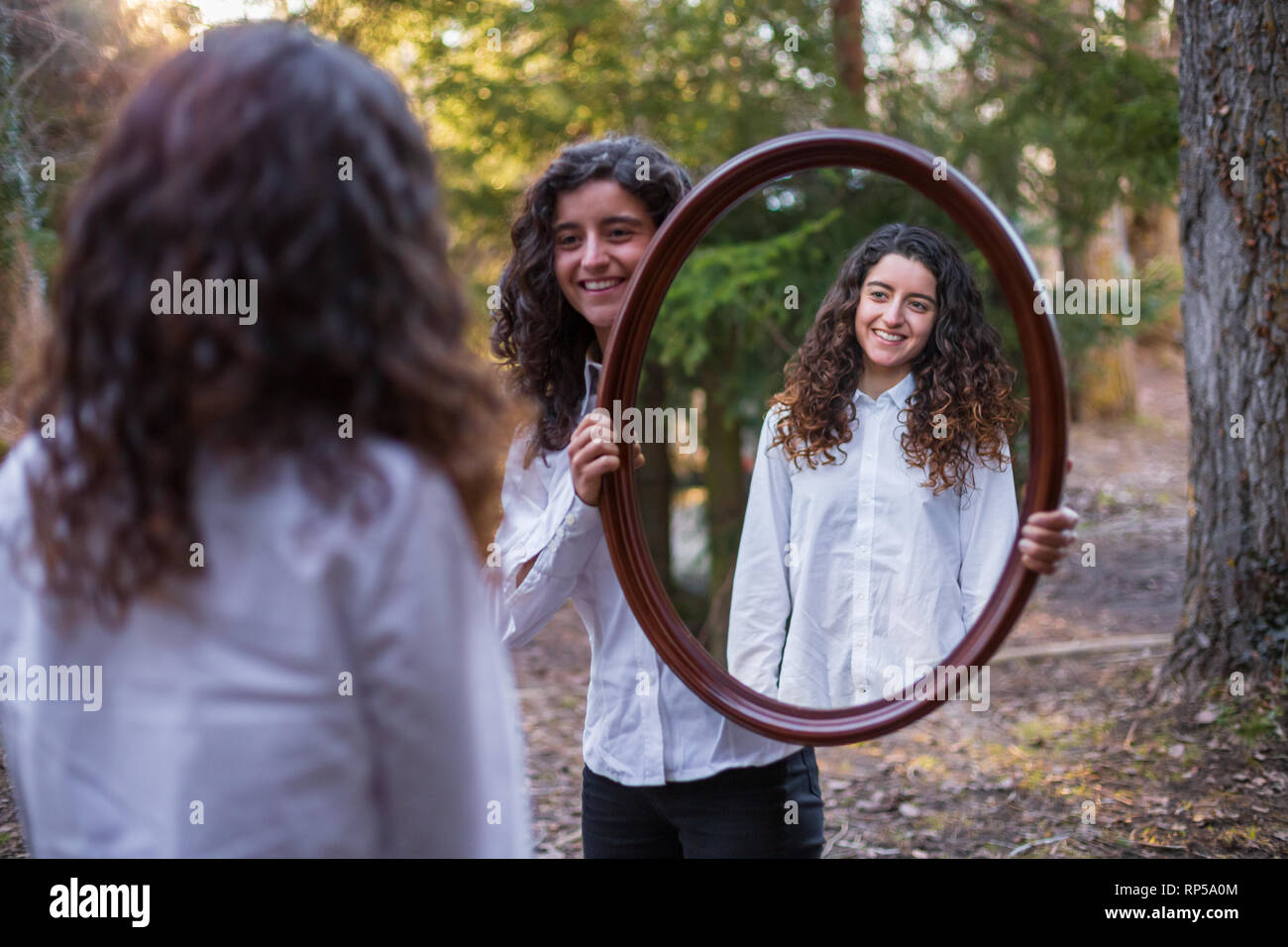 Cheerful young woman showing reflection of twin sister in autumn day in the forest Stock Photo