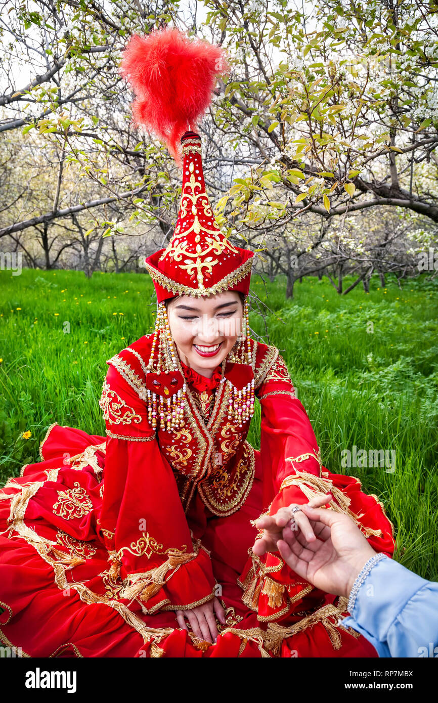 Happy woman in Kazakh red costume with wedding ring present of her boyfriend at spring garden in Kazakhstan. Proposal concept. Stock Photo