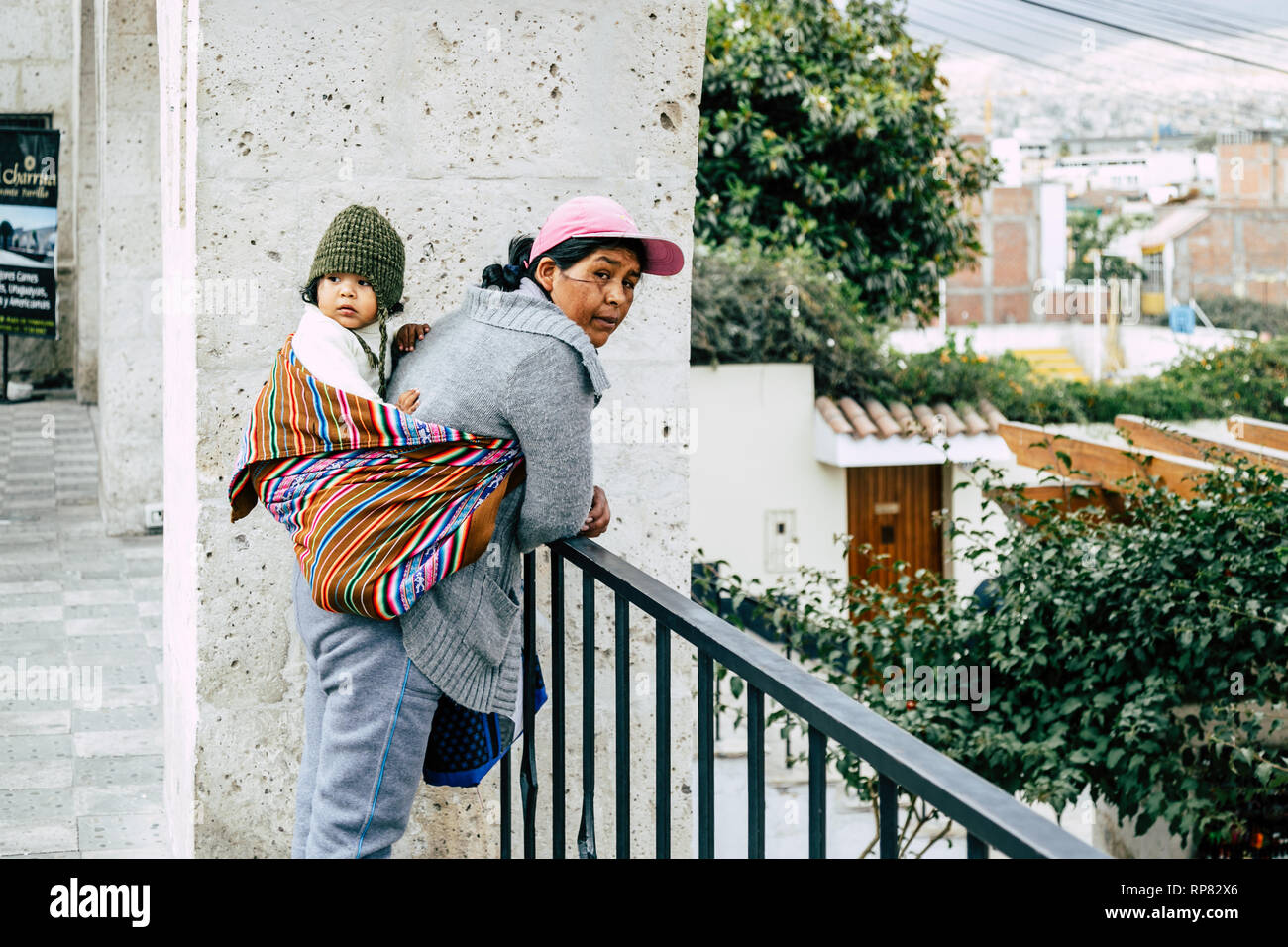 A mother leaning against a rail, waiting, while carrying her child on her back using a colourful manta in Arequipa (Peru). Stock Photo