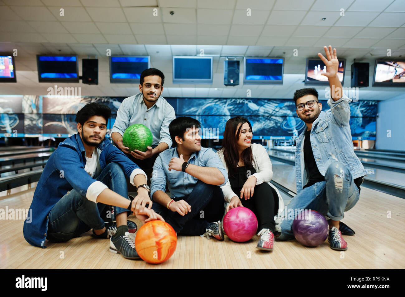 Group of five south asian peoples having rest and fun at bowling club, sitting on bowling alley with balls on hands. Stock Photo