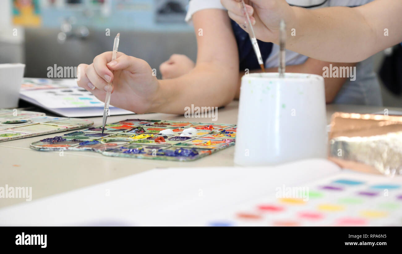 close up of student hands in visual art class holding a paint brush dipping in bright colours on a paint palette. Creative visual arts practical. Stock Photo