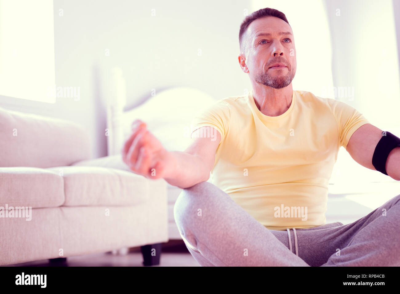 Man wearing grey trousers and yellow shirt sitting in lotus position meditating Stock Photo