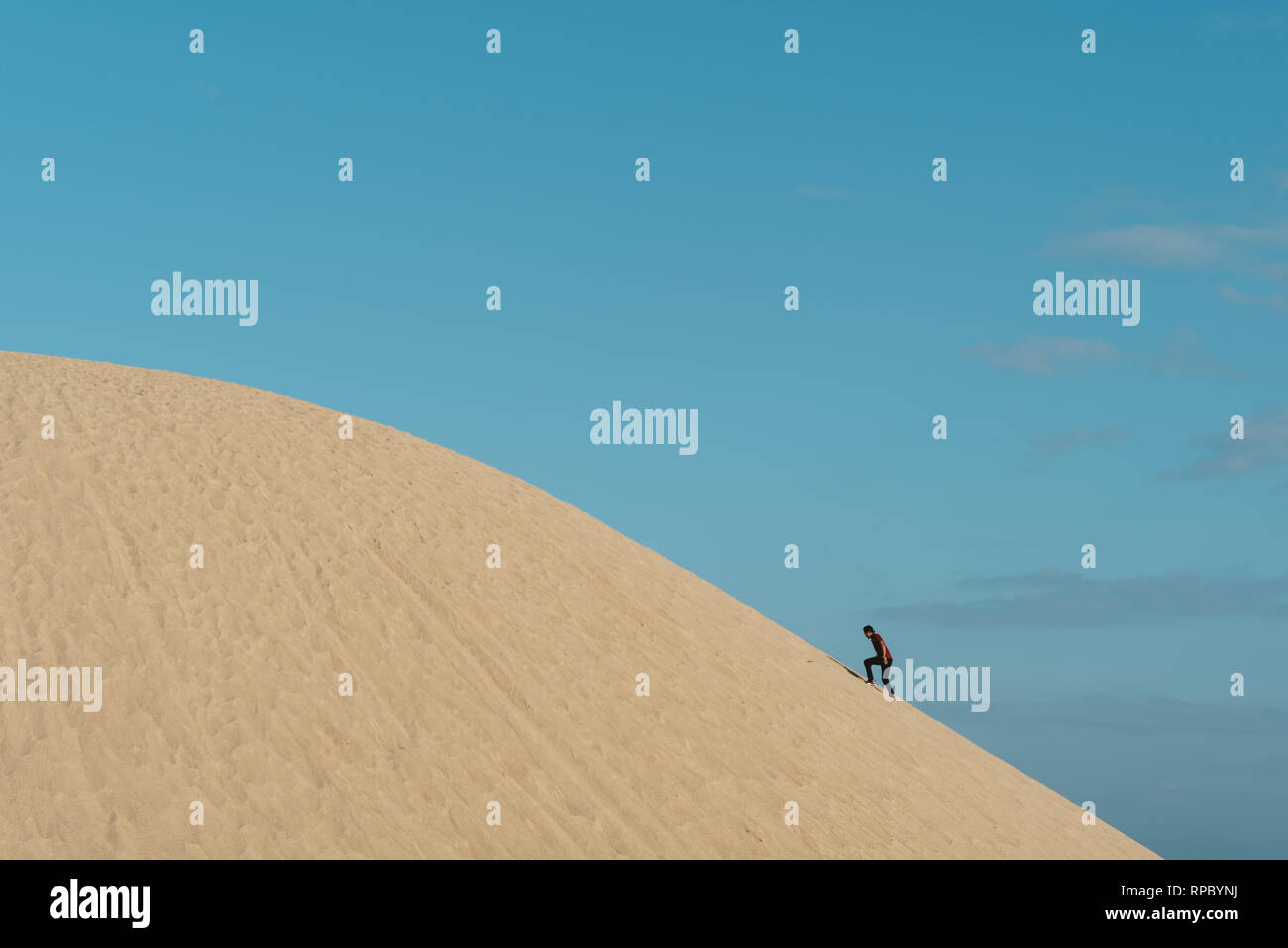 young adult asian male climbing a sand dune against blu sky background. concept of traveler, effort and nature Stock Photo