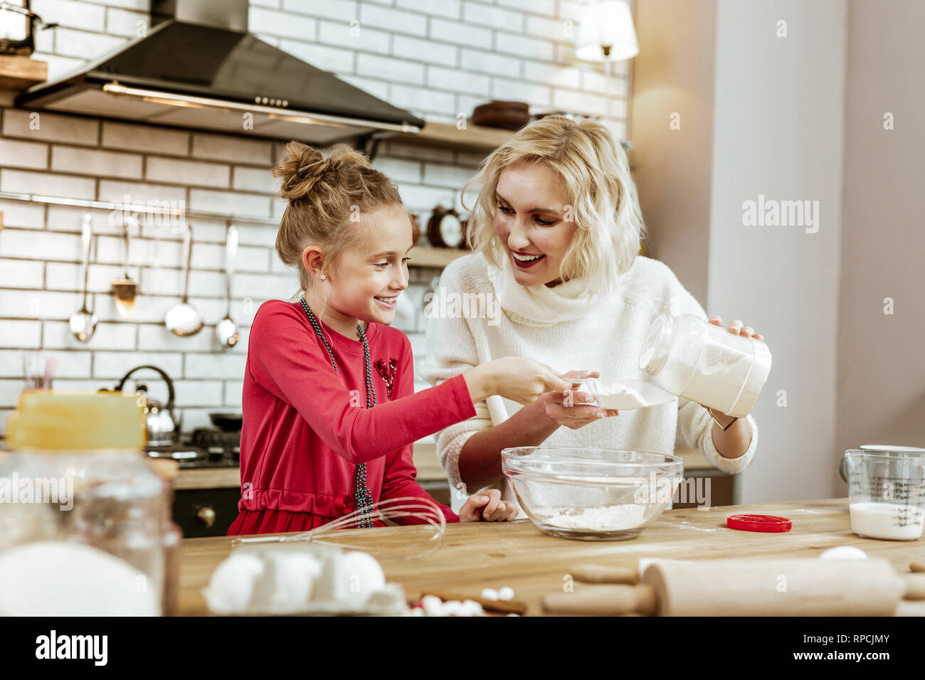 Light-haired beaming girl in red dress carrying measuring cup Stock Photo