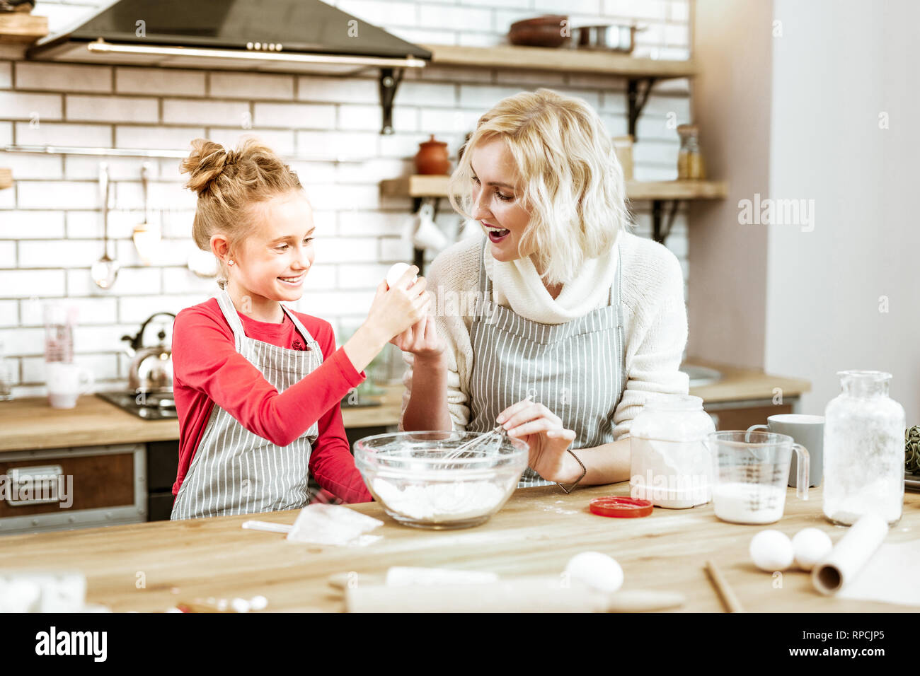 Beaming blonde mother in striped apron giving egg to curious daughter Stock Photo