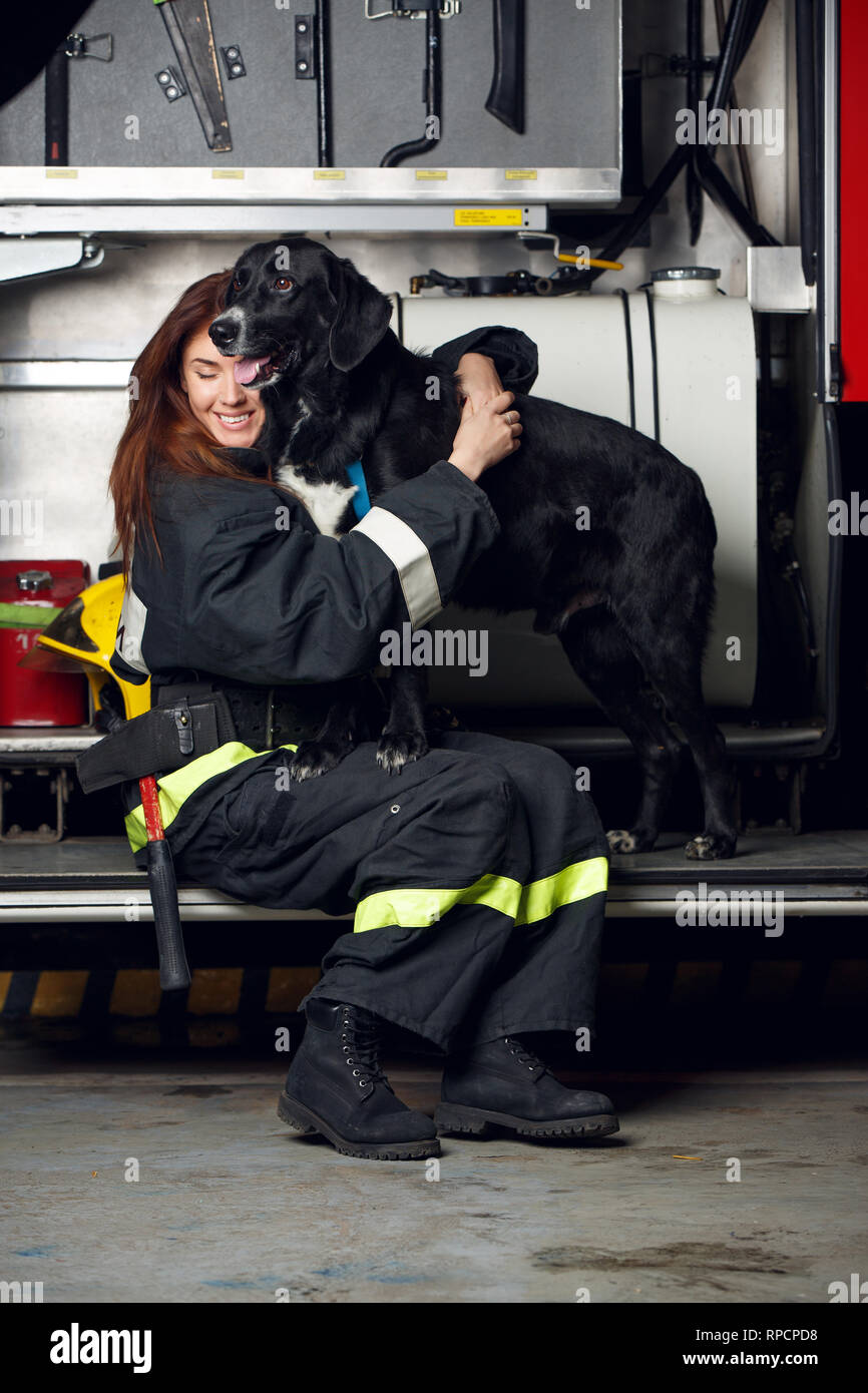 Portrait of young woman firefighter with black dog sitting on background of fire truck Stock Photo