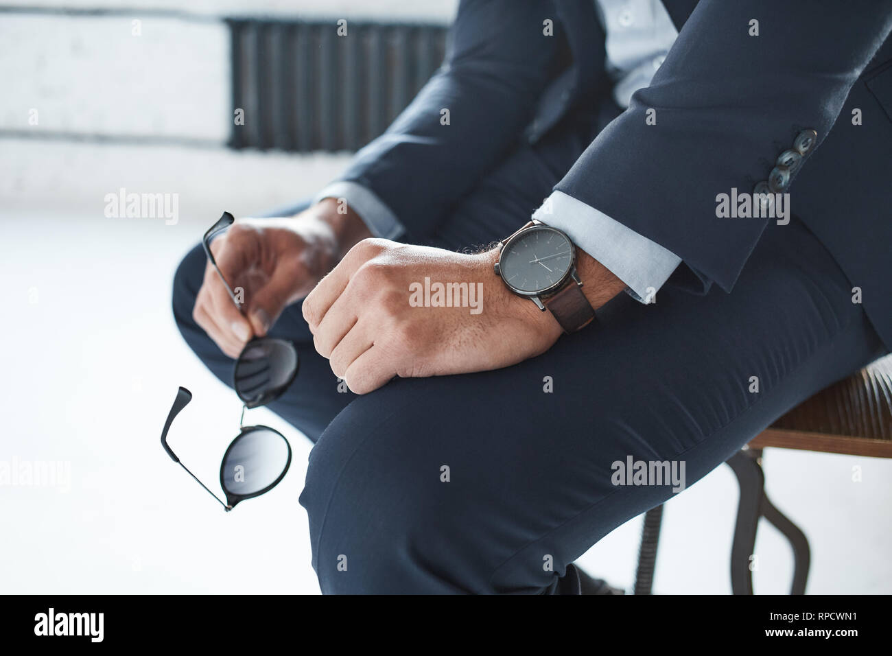 Close-up image of a stylish businessman who is sitting on the chair with branded watch on his hand and holding glasses. Business look at office Stock Photo