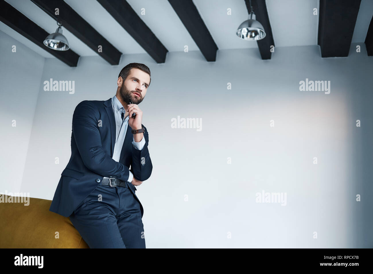 Looking just perfect. Young bearded businessman in a stylish dark suit is thinking about successful future in his modern office. Fashion look. Business style. Stock Photo