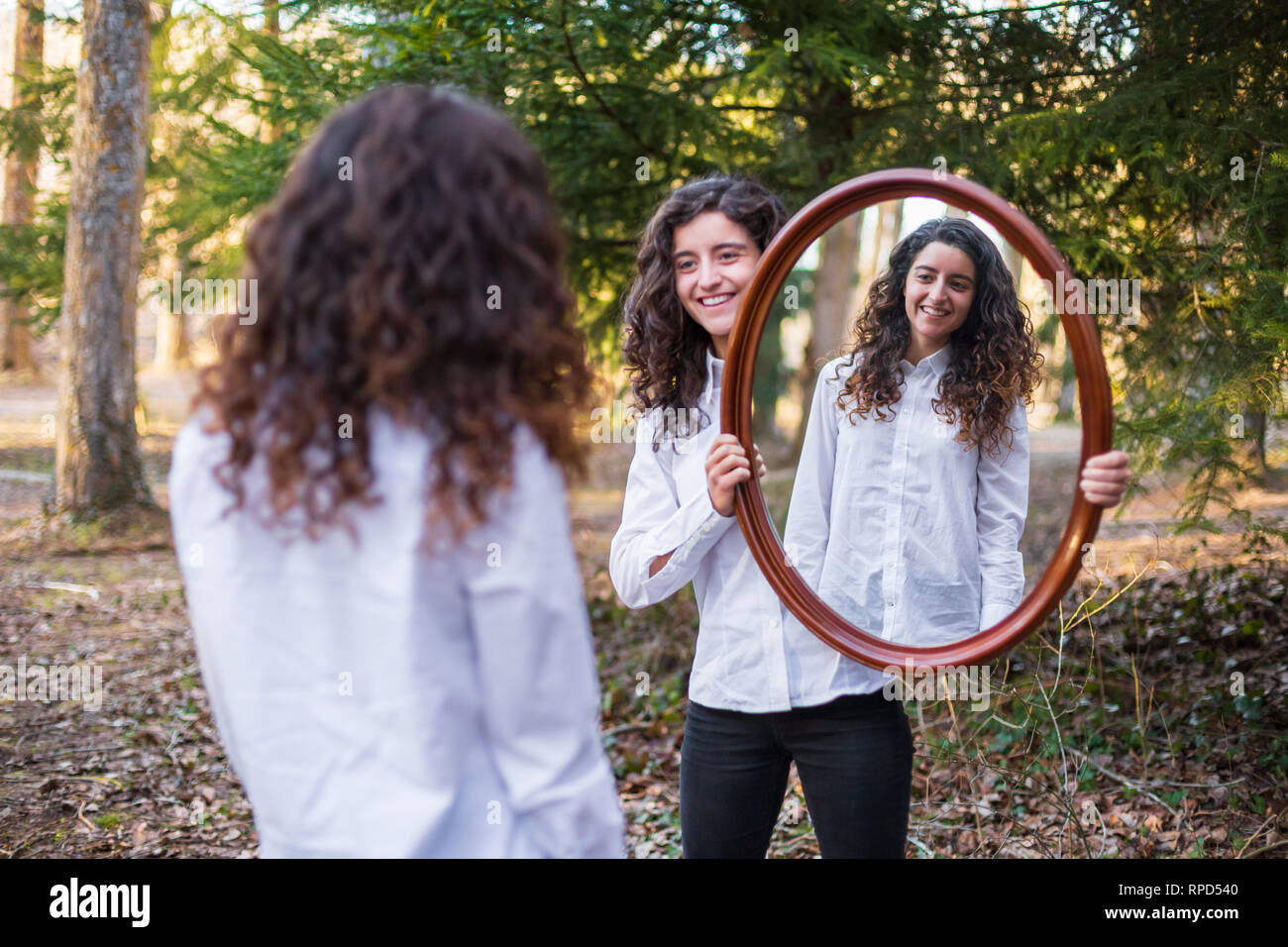 Cheerful young woman showing reflection of twin sister in autumn day in the forest Stock Photo