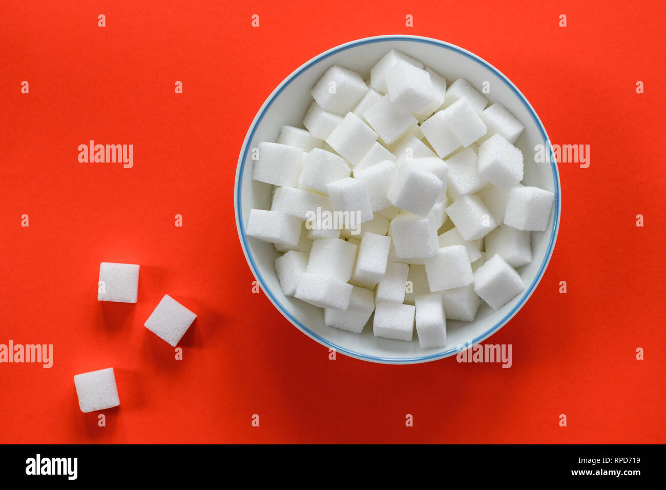 Top view of a white ceramic bowl filled with sugar cubes isolated on a red background. A few sugar cubes are spread on the table. Stock Photo