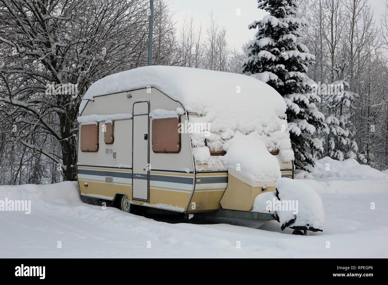 Caravan trailer in a parking lot next to a forest covered with snow during winter in Europe. The trailer is yellow and grey colored. Stock Photo