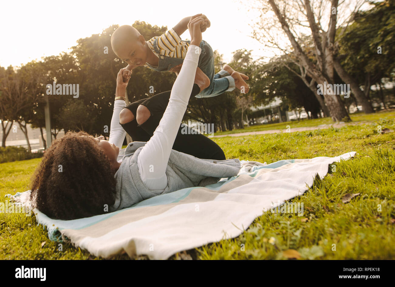 Mother playing with his son at park. African woman lying on plaid at the park and raising the boy on her feet. Stock Photo