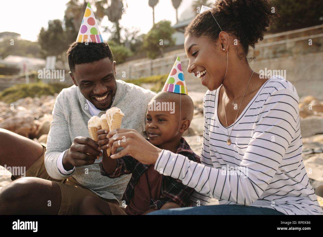 African family with party hats eating ice cream on the beach. Man and woman with son enjoying eating ice-cream. Family enjoying son's birthday with ic Stock Photo