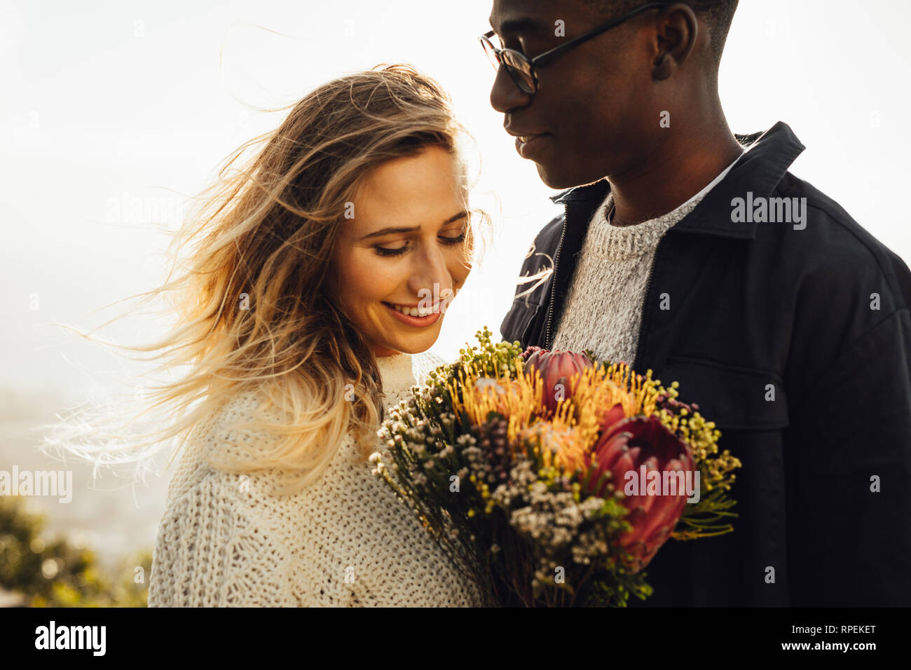 Young man looking at his girlfriend with flowers outdoors. Interracial couple standing together with bunch of flowers. Stock Photo