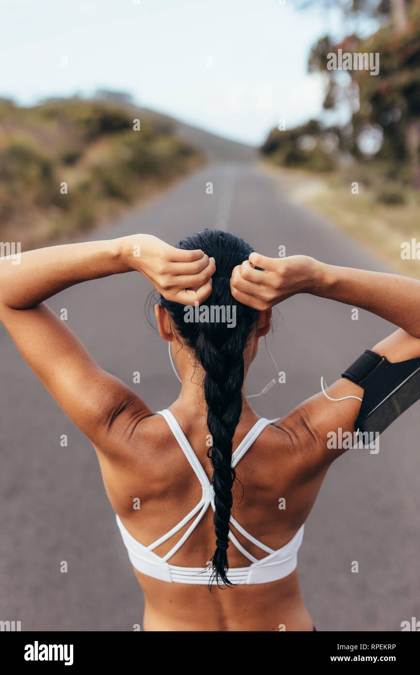 Rear view shot of fit female athlete tying hair before her workout. Young woman getting ready for training outdoors on an empty road. Stock Photo